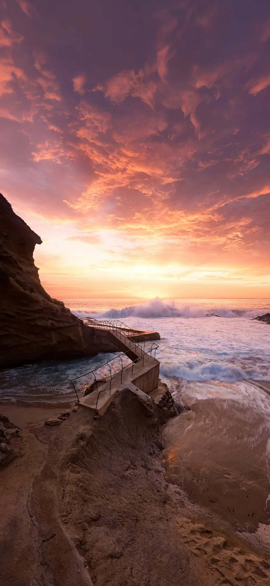 Sunset over crashing waves and rocky walkway at Laguna Beach, United States, featured as a dramatic coastal Phone Wallpaper.