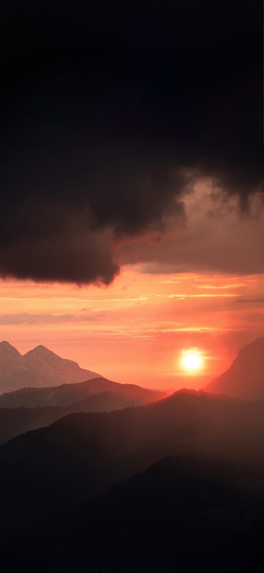 Fiery sunset over layered mountain peaks at Col des Aravis in La Clusaz, France, with dramatic clouds and glowing horizon – Phone Wallpaper.