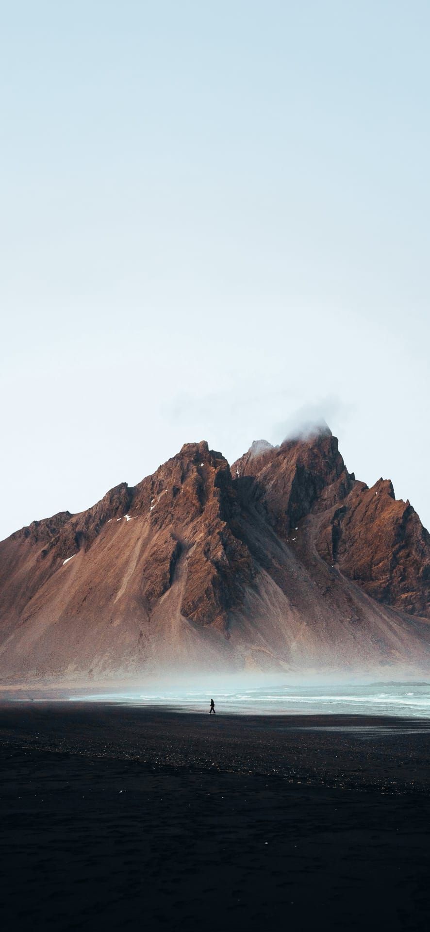Person walking along a black sand beach with misty coastal mountains in the background, perfect for dramatic nature Phone Wallpaper.